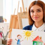 Злата Зайферт учитель ИЗО в Москве держит детский рисунок с солнцем и домом