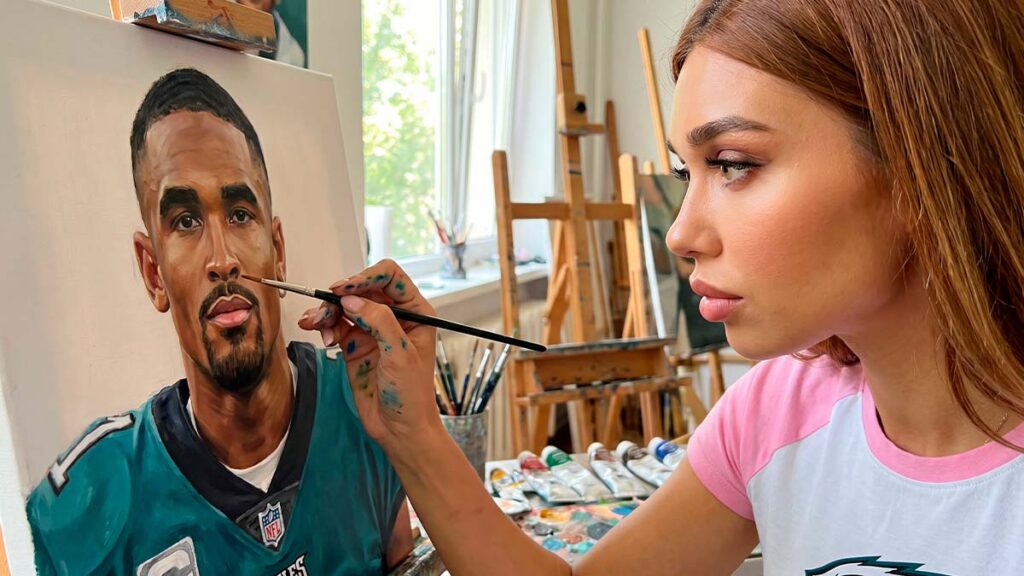 Зайферт Злата нарисовала портрет Jalen Hurts 2 Зайферт Злата в бело-розовой футболке с испачканными краской руками прорисовывает кистью лицо на портрете спортсмена в форме «Филадельфия Иглз».